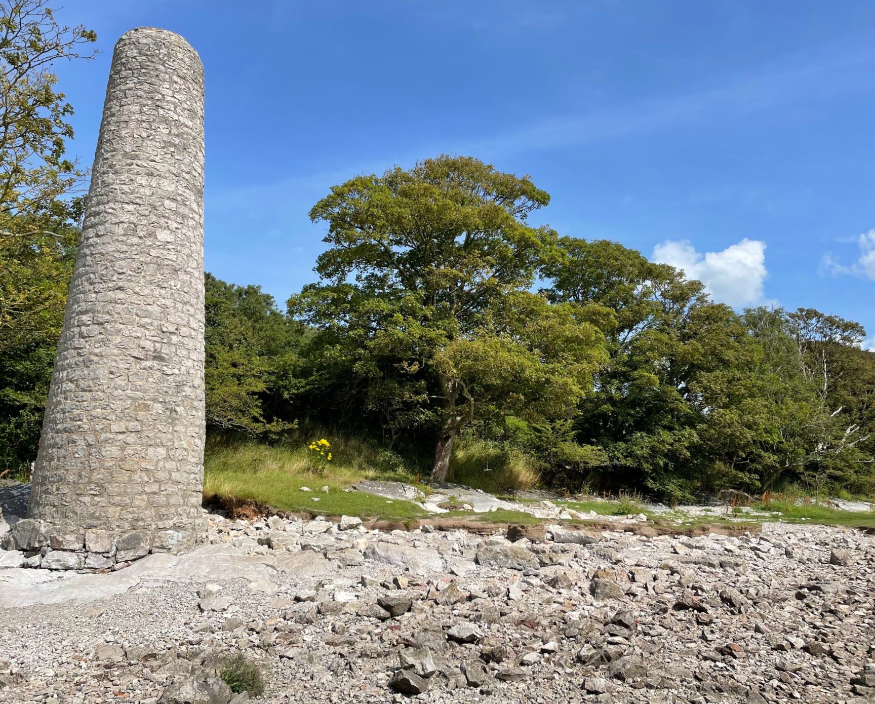 A limestone cylindrical tower on a rocky foreshore with trees in the background.
