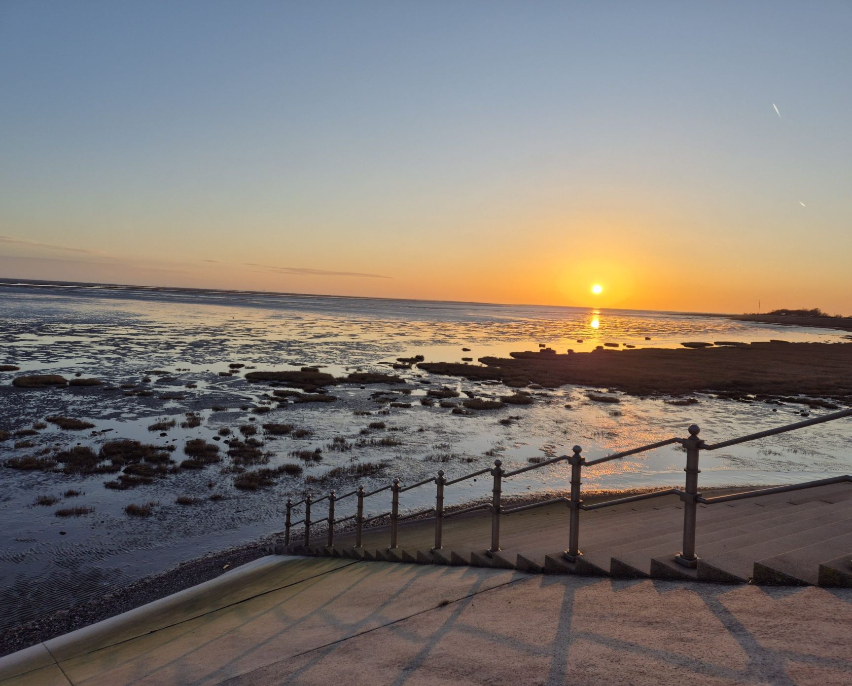A wet beach at sunset with tufts of spartina grass, railings in the foreground.